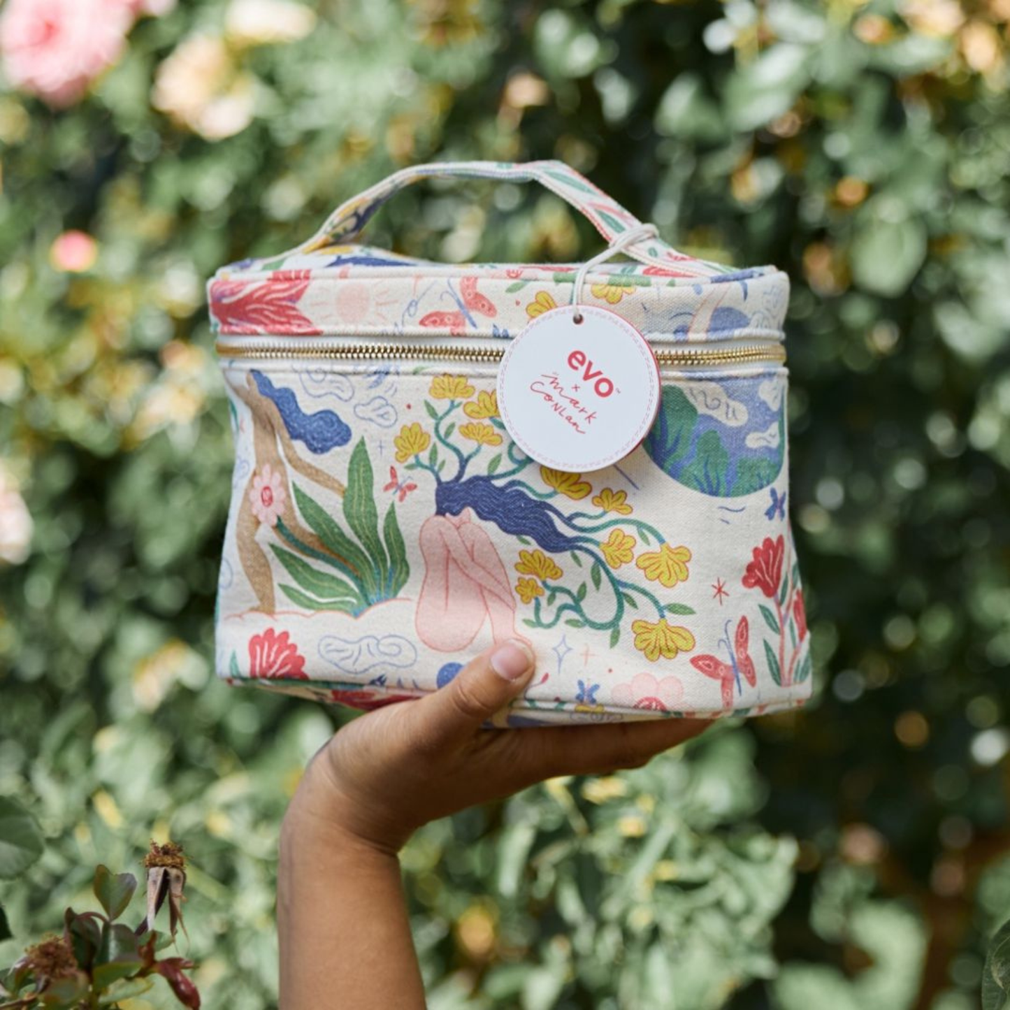Hand holding a Mark Conlon patterned makeup bag with 'evo' brand tag against a green foliage background