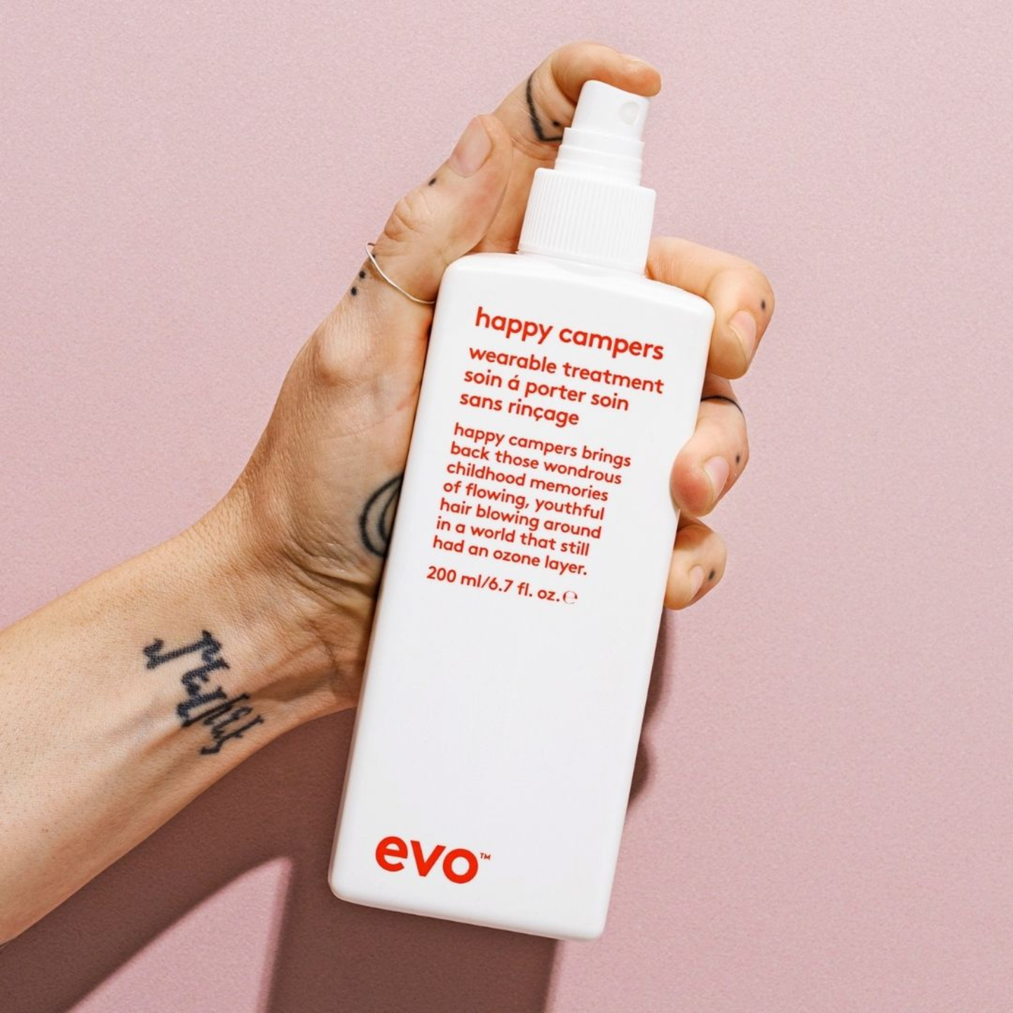 Hand holding a bottle of Happy Campers Wearable Treatment by evo against a pink background