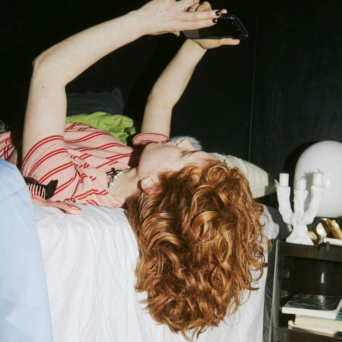 Woman with wavy red hair laying on her bed looking at her phone