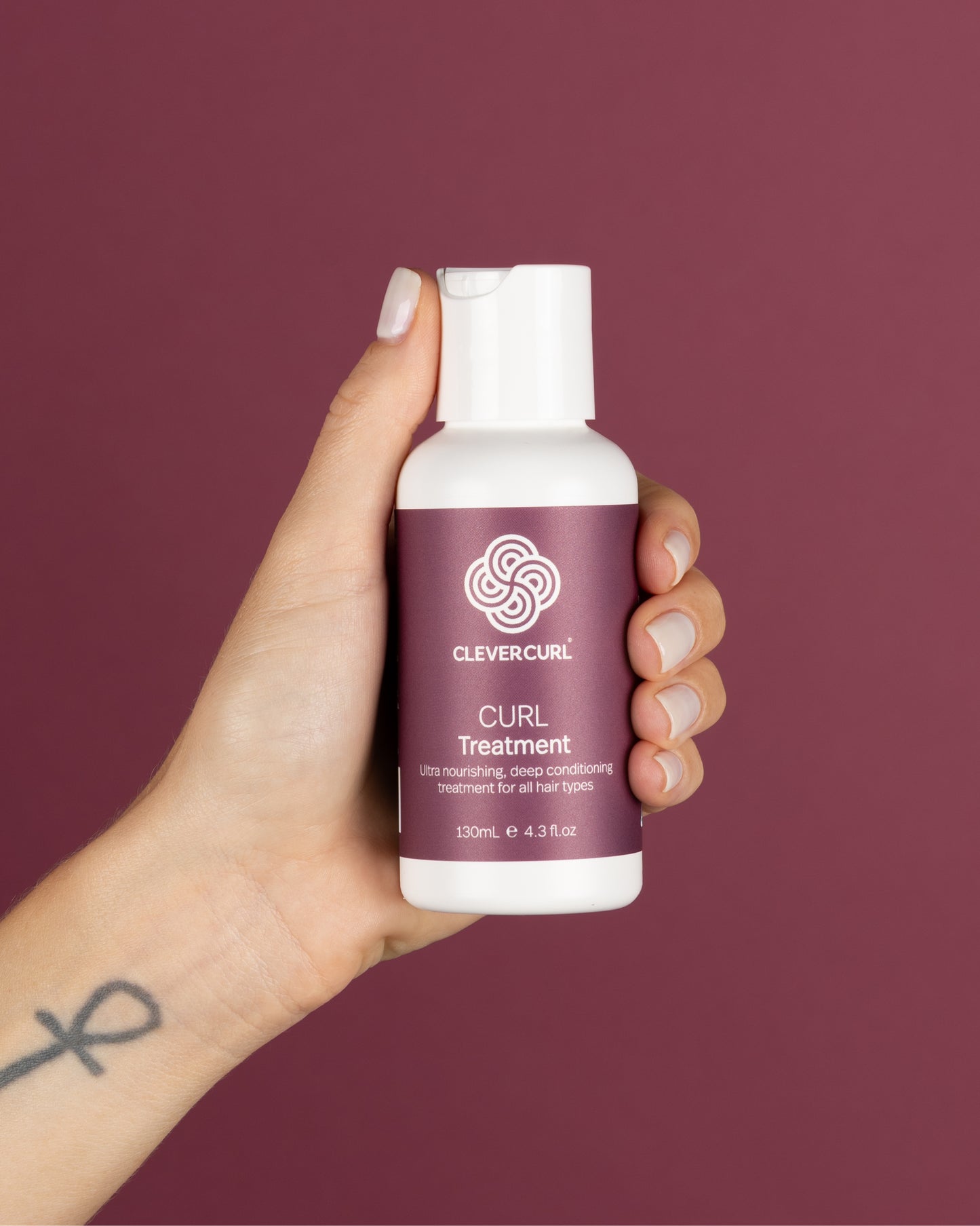 Hand holding a bottle of Clevercurl Curl Treatment 130ml against a maroon background