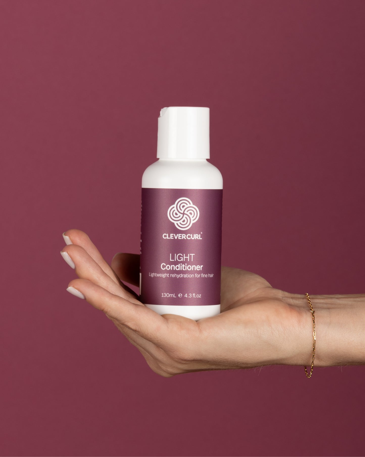 Hand holding a bottle of Clever Curl Light Conditioner 130ml against a maroon background