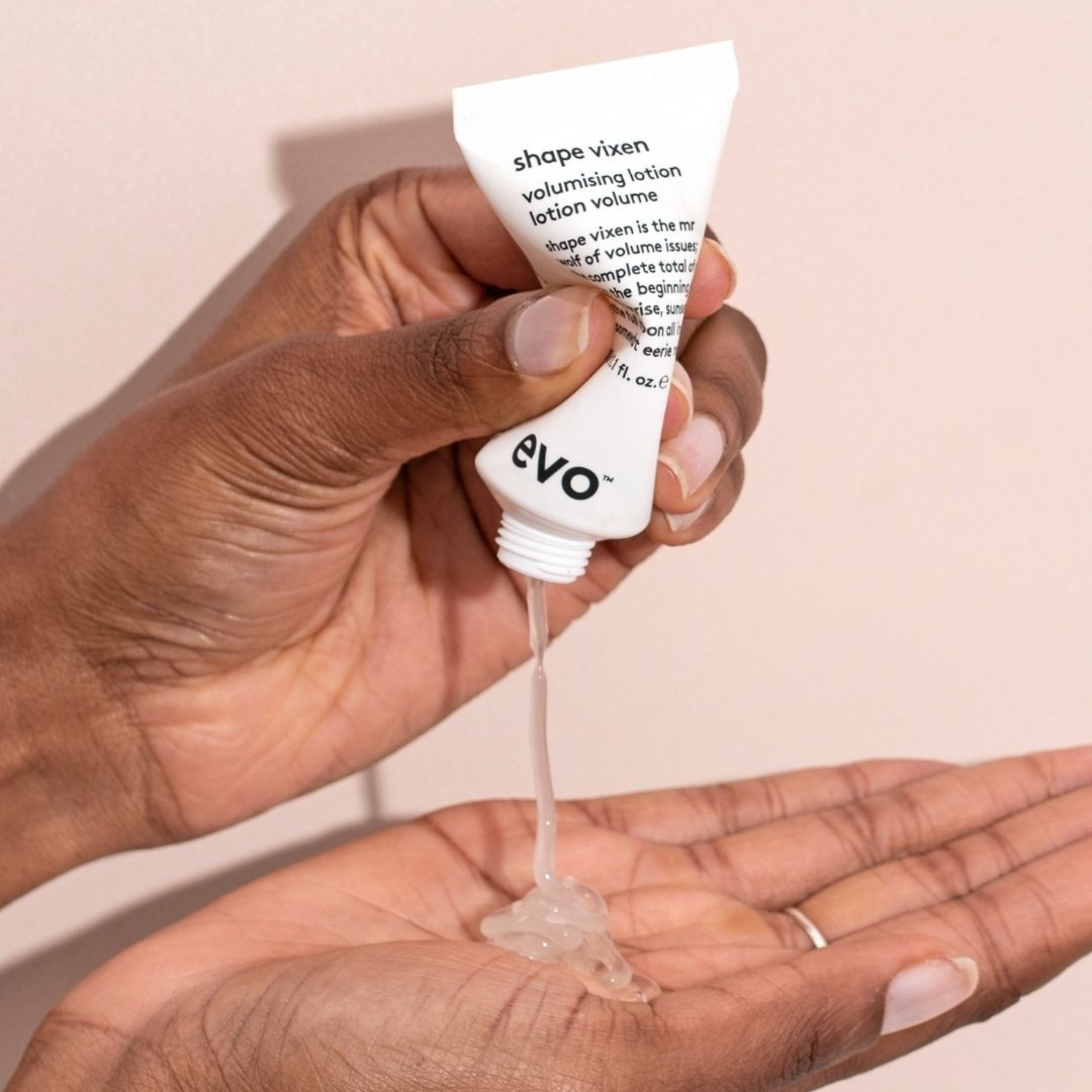 Hand holding a tube of evo shape vixen volumizing lotion with liquid dripping onto a palm against a beige background.