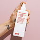 Hand holding a bottle of Happy Campers Wearable Treatment by evo against a pink background